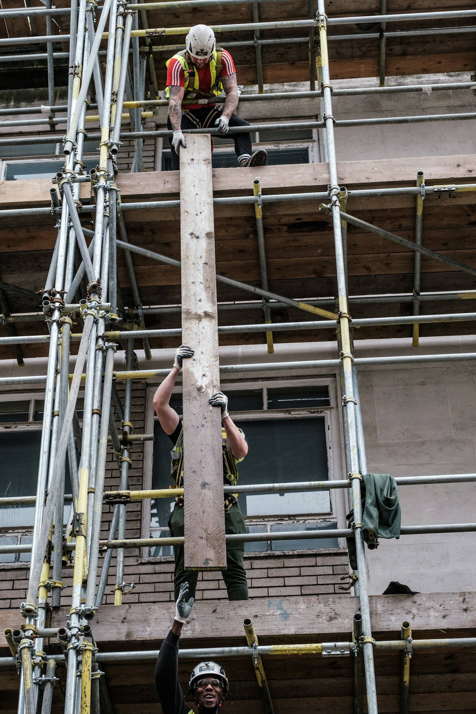 Two construction workers on scaffolding lifting a wooden beam outside a brick building.
