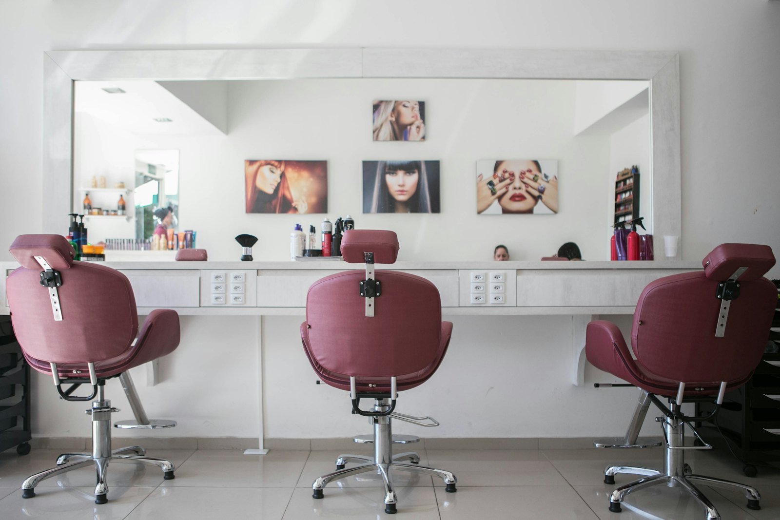 An independent hair salon interior with three pink-leather chairs, a long mirror, and bottles on a white shelf.